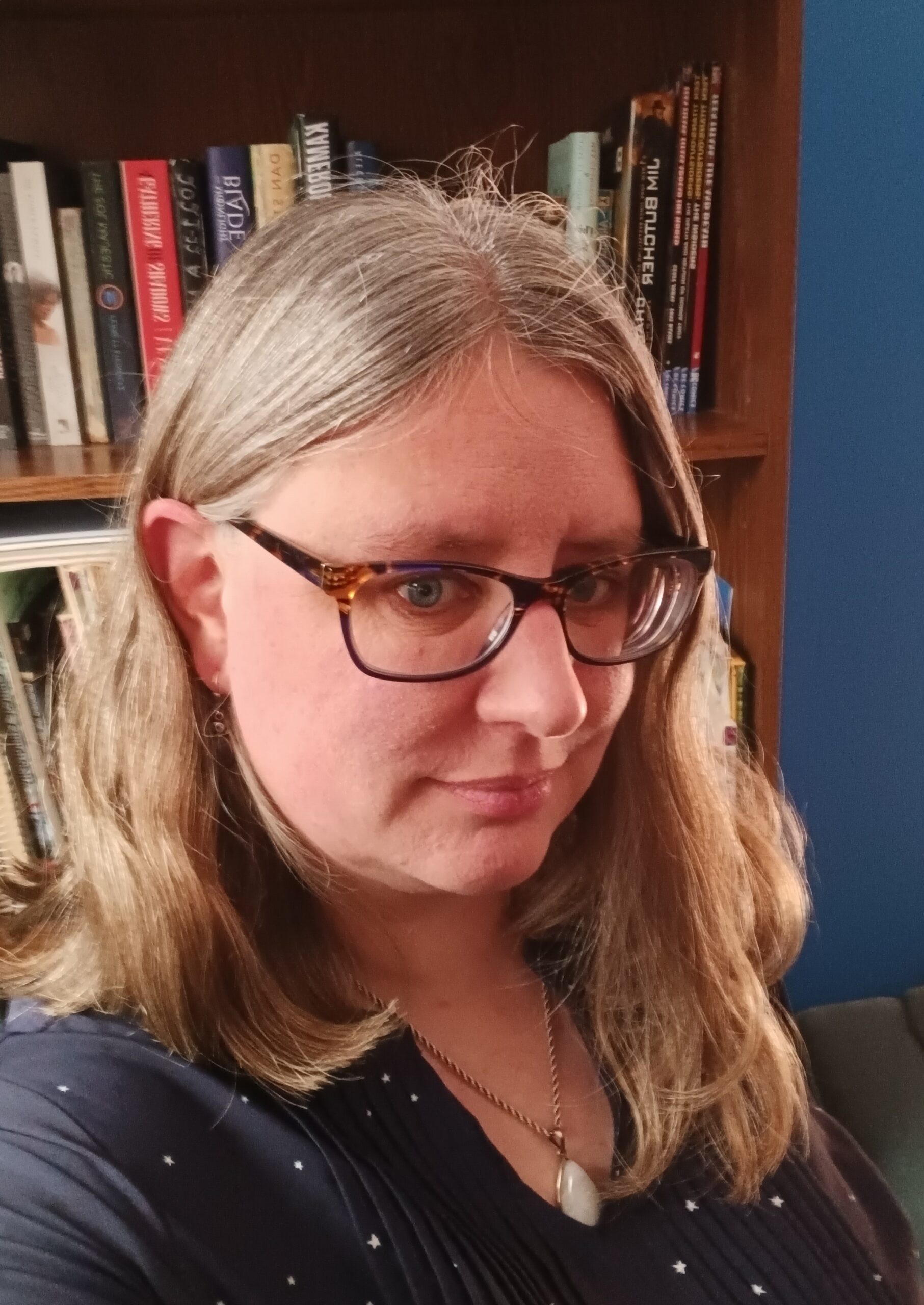 A middle aged white women with greying hair and glasses looking at the camera slightly smiling with a bookshelf in the backgound